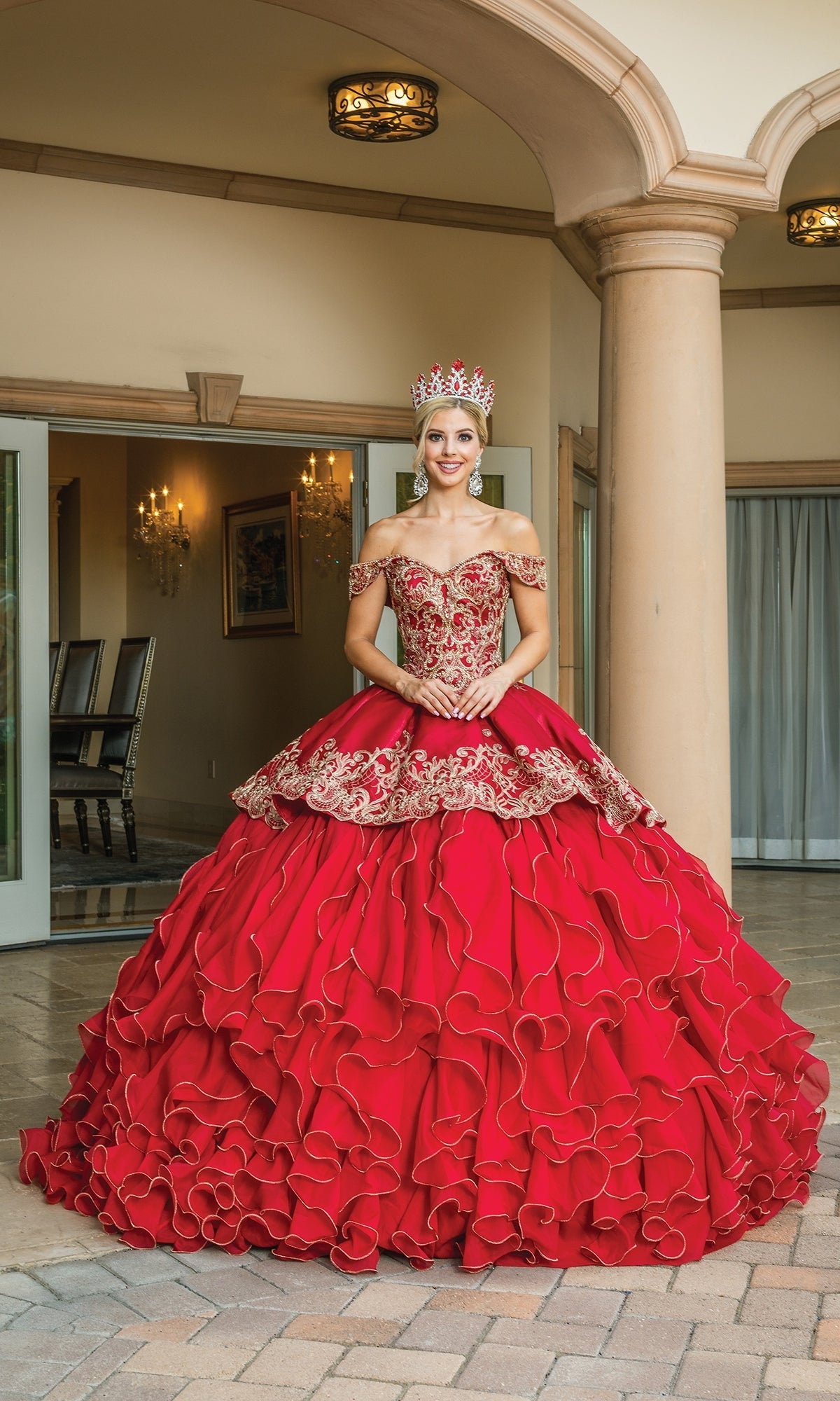 A woman in the Quinceanera Dress 1599 by Dancing Queen—an ornate red ball gown with gold embroidery, a matching crown, and off-the-shoulder ruffle sleeves—stands before a building with columns and open doors.